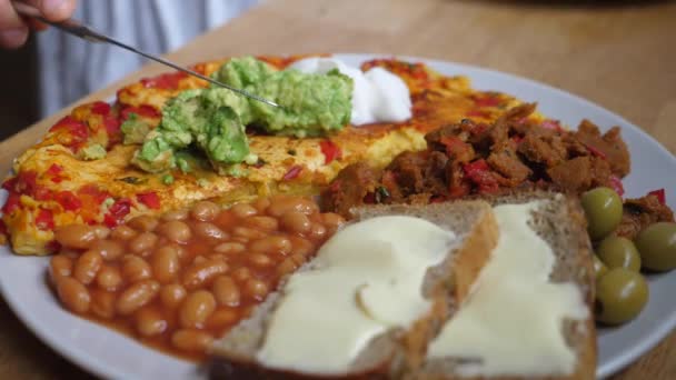 Gesundes, nährstoffreiches Frühstück zu Hause. Avocado-Smash auf Tofu-Gerangel verteilen. Mahlzeit auf weißem Teller serviert auf Holztisch — Stockvideo