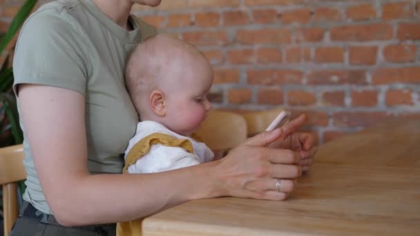 Bayi Kaukasia lucu bermain di smartphone sambil duduk di pangkuan ibunya. Teknologi dan anak-anak — Stok Video