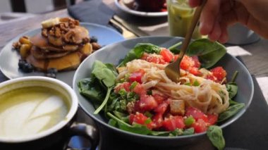 Yapraklı ve sebzeli makarna salatası yiyorum. Dışarıda sağlıklı ve lezzetli bir vejetaryen kahvaltısı. Arka planda krep ve içecekler yığını.