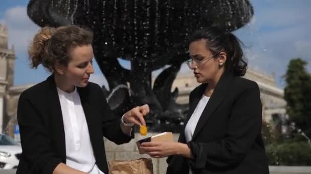 Дві жінки-співробітниці, насолоджуючись обідом, відпочивають на свіжому повітрі, забираючи свою частку біля фонтану. Підтримка здорового робочого місця — стокове відео
