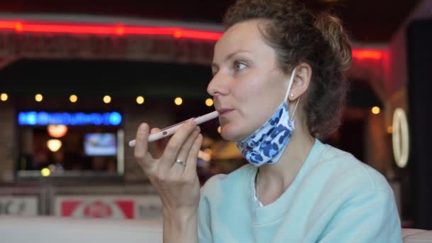 Kaukasisches Mädchen, das mit ihrem Smartphone eine Audiobotschaft aufzeichnet, mit Gesichtsmaske, die am Kinn in einem Kaffeehaus hängt. Zweite Welle der Covid-19-Pandemie . — Stockvideo