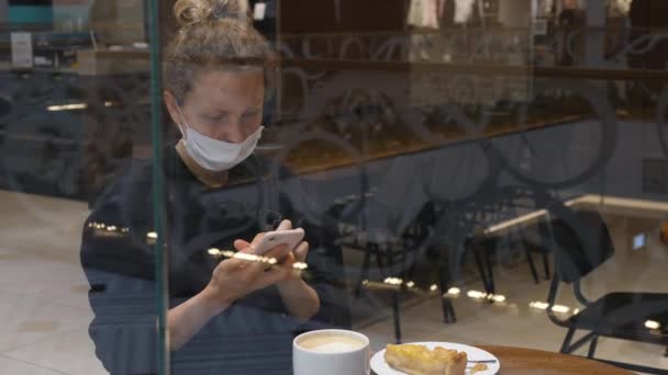 Menina no local do café usando seu smartphone no coffee break com cofffee e torta durante a pandemia de covid-19. — Vídeo de Stock