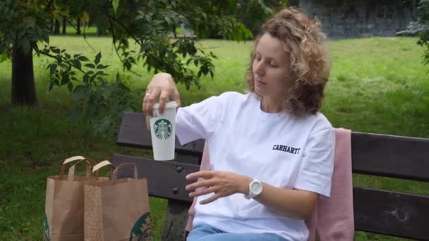 Vit tjej i parken sitter på bänken och njuter av sin Starbucks-drink. Warszawa-Polen-2020 — Stockvideo