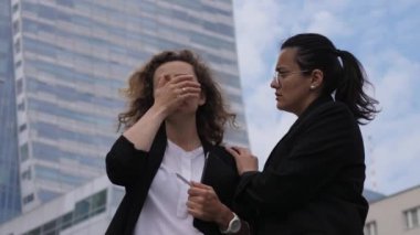 Esmer şirket çalışanı, stresli sarışın meslektaşını gökdelenin önünde neşelendiriyor.