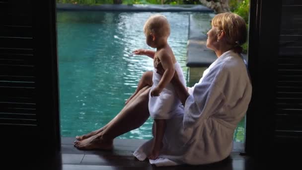 Mormor satt på kanten av poolen med sitt barnbarn i en vit morgonrock. Lycklig familj på semester — Stockvideo