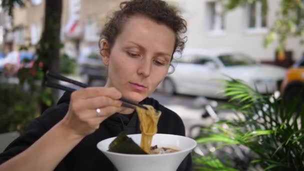 Vue latérale de la femme affamée mangeant des nouilles miso riches en légumes avec des baguettes dans le parc. Santé végétalien asiatique street food — Video