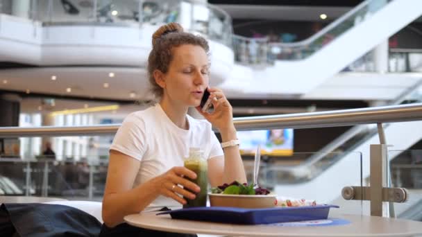 Обідня перерва бізнес-леді. Молода жінка розмовляє по телефону, посміхається і п'є свою зелену смужку під час обіду на продовольчому майданчику торгового центру. Ескалатори на фоні — стокове відео