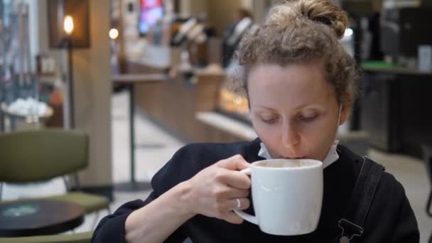 Fiatal kaukázusi nő leveszi az arcmaszkot, hogy igyon egy korty forró italt a kávézóban a kovid-19-es járvány idején.. — Stock videók