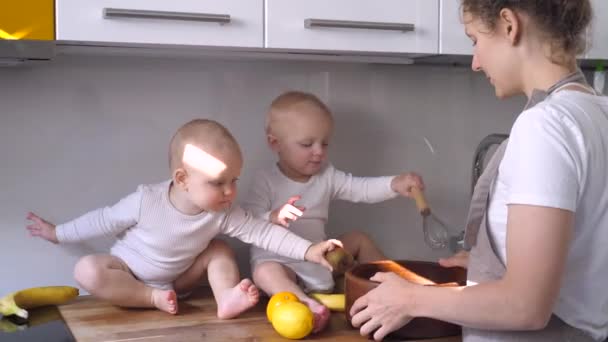 Concepto familiar feliz. Preciosas hermanas gemelas juegan con frutas en la cocina mientras la madre está cocinando comida saludable — Vídeos de Stock