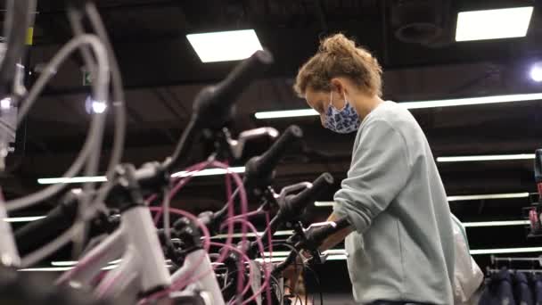 Elegir una bicicleta. Mujer joven con mascarilla protectora comprueba los frenos en las bicicletas en la tienda de ciclistas — Vídeo de stock