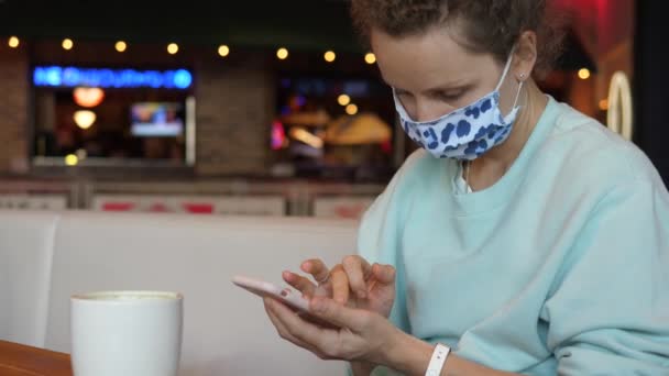 Ung kvinna bär ansiktsmask med hjälp av sin smartphone på kaffe plats på sin kaffepaus. Andra vågen av covid-19 pandemi . — Stockvideo