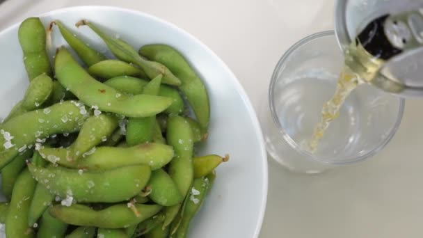 Ovanifrån av hälla läsk från burk till glas. Edamame bönor i en skål. Veganska japanska snacks — Stockvideo