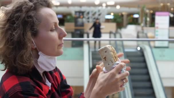 Manger dans les lieux publics pendant l'épidémie de covidé-19. Vue latérale de la femme blanche enlever masque réutilisable pour mordre enveloppement végétarien dans le centre commercial. — Video