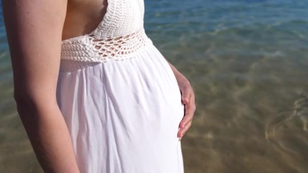 Zwangere vrouw draagt witte jurk op het strand aan de zee. — Stockvideo