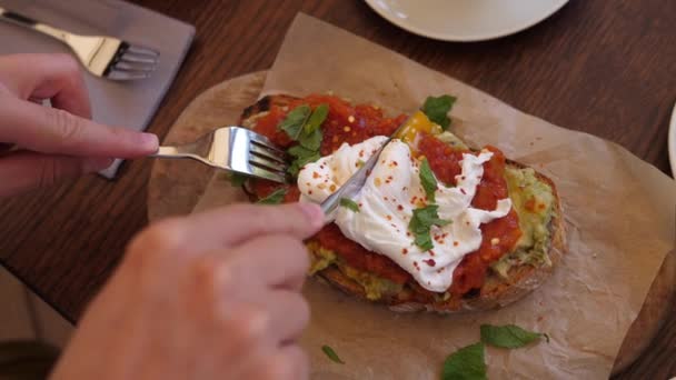 Spécialité petit déjeuner méditerranéen équilibré. Vue du dessus du couteau et de la fourchette coupés en parfait œuf poché sur un pain grillé à l'avocat avec tartinade de tomates servi à bord — Video