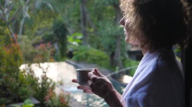 Bali 'de bir villada sabah. Bir fincan sıcak içecekle tropik manzaranın tadını çıkaran bir kadın.