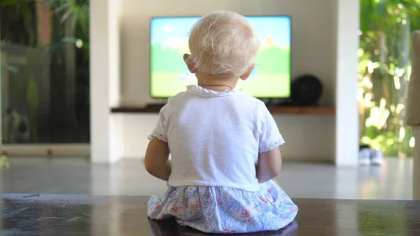 Back view of a little girl sitting on the floor watching the tv