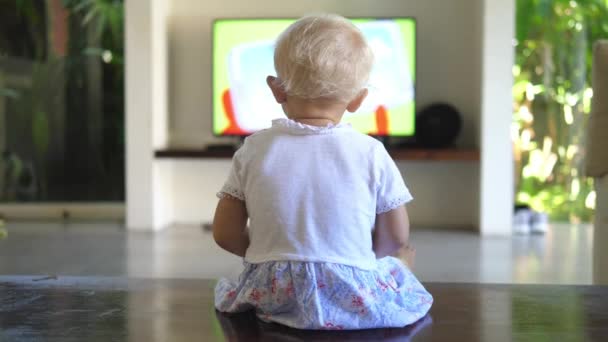 Bakspeilet av en liten jente som sitter på gulvet og ser på tv – stockvideo