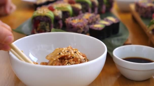 Kimchi von Hand aus einer kleinen weißen Porzellanschüssel pflücken. Sets von buntem Sushi im Hintergrund. Asiatisches Streetfood-Konzept — Stockvideo