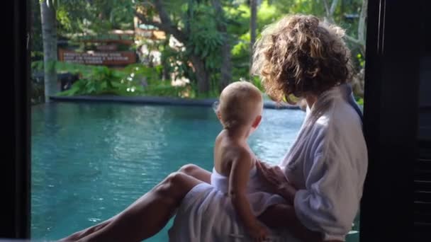 Вид сзади матери с ребенком на коленях, сидящей и смотрящей на бассейн. Оба одеты в белые халаты — стоковое видео