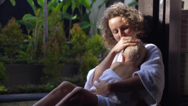 Bornozlu genç beyaz bir anne, havuz kenarında bebeğine sarılıyor. Aile tatili kavramı — Stok video