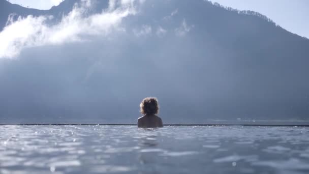 Vista trasera de una silueta de una mujer saltando de las aguas termales naturales de Batur. Batur montaña en la parte posterior — Vídeo de stock