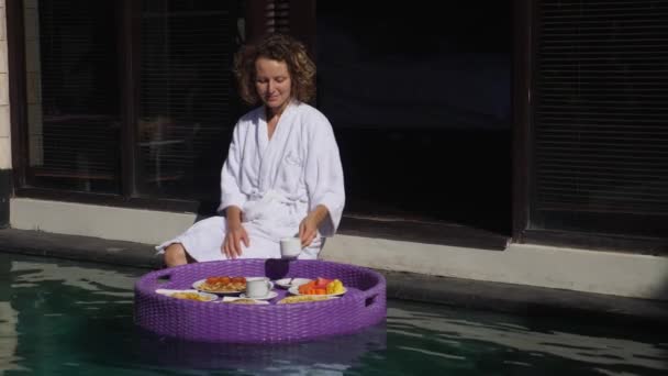 Concepto de desayuno flotante. Mañana de una mujer rubia en un albornoz en un lujoso resort. — Vídeos de Stock