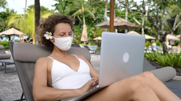Zdalna praca na plaży podczas wirusa korony. Biała kobieta w masce na twarzy z laptopem na leżaku — Zdjęcie stockowe