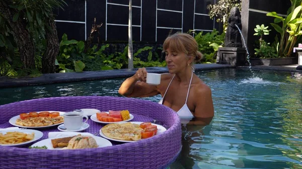 En medelålders kvinna i en swimmingpool som äter en flytande frukost. Livet efter pensioneringen — Stockfoto