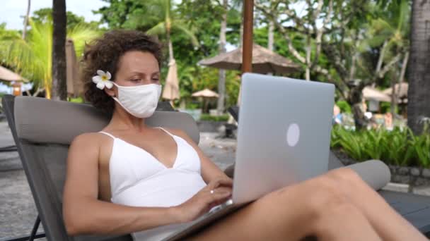 Trabalho remoto na praia durante o vírus corona. Mulher caucasiana em uma máscara facial com um laptop em uma espreguiçadeira — Vídeo de Stock