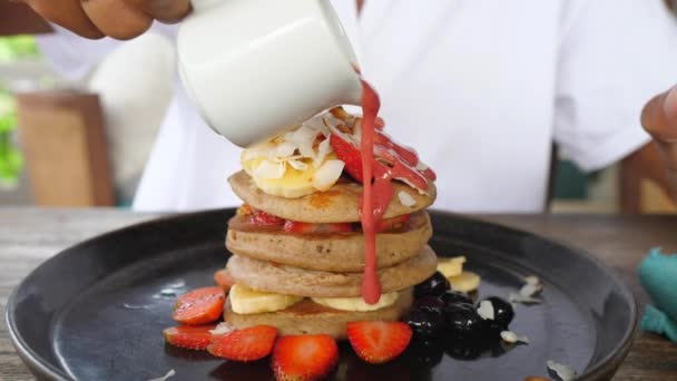 Close-up van de hand gieten van een aardbeisaus op een stapel veganistische pannenkoeken versierd met bessen — Stockvideo