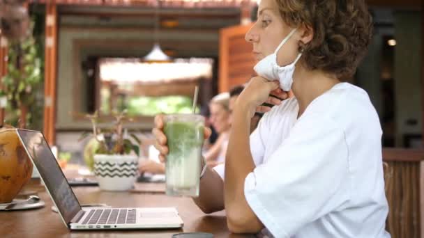 Remote work during a covid-19 pandemic. A young woman in a face mask working at a coffee place, takes off the face mask and sips matcha tea latte — Stock Video
