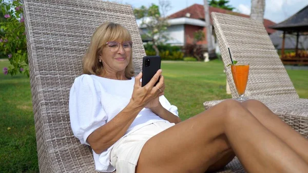 Kvinne over 50 på solseng smiler til smarttelefonen sin mens video ringer med elskede. Forbli tilkoplet i en rikstelefon. – stockfoto