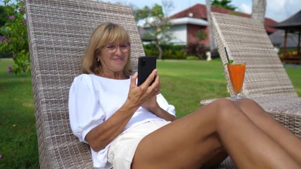 Woman over 50 on a sunbed smiling at her smartphone while video calling with beloved ones. Staying connected on a long distance. — Stock Video