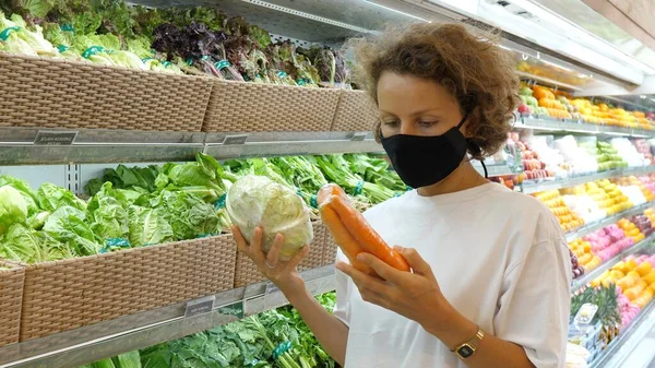 A woman in a face mask comparing carefully pre packed vegetables and chooses the best ones. Grocery shopping during covid-19 pandemic.