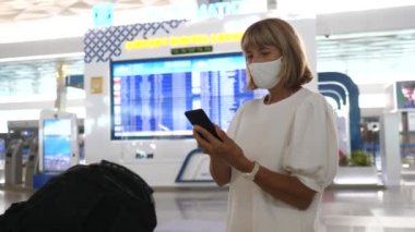 Yüzü maskeli orta yaşlı bir kadın. Telefonunu kontrol ediyor. Havaalanının kalkış panosunda duruyor. 