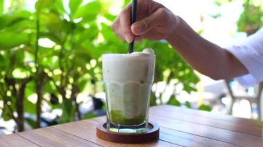 Healthy delicious refreshing plant based drinks on a sunny day. Close up of hand stirring matcha ice tea latte with almond milk.
