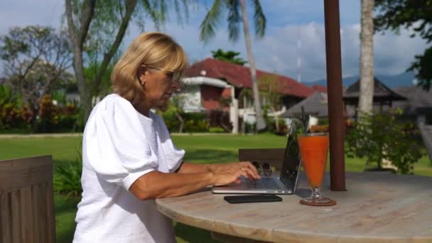 Trabalhando remotamente em feriados tropicais. Empreendedor de meia idade senhora chefe trabalhando em um laptop durante as férias em uma ilha — Vídeo de Stock