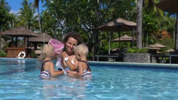 Tips til, hvordan man holder børnene beskæftiget på ferie. Mor og tvilling døtre i en pool med spanden og andet legetøj. – Stock-video