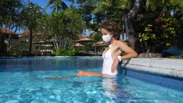 Coronavirus-Einschränkungen: Eine junge weiße Frau trägt eine Gesichtsmaske, während sie allein im Schwimmbad chillt. Sonniger Tag auf einer tropischen Insel — Stockvideo