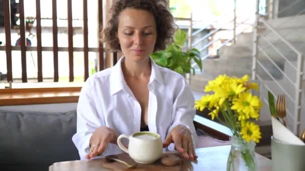 Vrolijk blond meisje genieten van haar koffiepauze van matcha thee latte en een plantaardige koekje. — Stockvideo