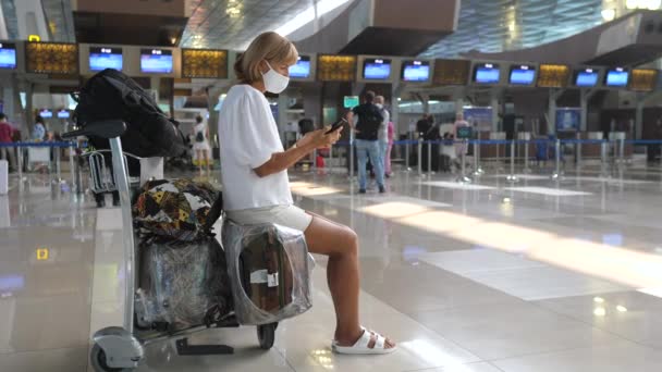 Utazás a covid-19-es járvány idején. Egy nő arcmaszkot visel a reptéren, a telefonját nézi. — Stock videók