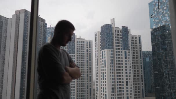 Social Isolation concept. Caucasian man with crossing arms looking at the buildings outside the window — Stock Video