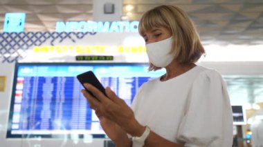 Covid-19 salgını sırasında seyahat etmek. Orta yaşlı, maske takan sarışın kadın çıkış panosunda kapı numarasını arıyor.. 