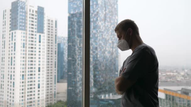 Aislamiento social que conduce a la soledad crónica y al aburrimiento. Hombre en la cara máscara de pie con los brazos cruzados por la ventana en una megápolis — Vídeos de Stock