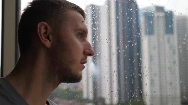 Efeitos de quarentena no bem-estar físico e mental. Vista lateral de um homem colocando a cabeça contra a janela em um dia chuvoso — Vídeo de Stock