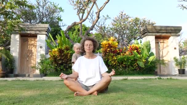 Genç anne Lotus pozisyonunda oturarak sakin kalmaya çalışıyor. Bu arada küçük bebeği onu rahatsız ediyor. — Stok video