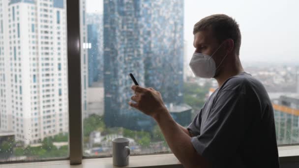 Занимаешься во время карантина. Человек в маске для лица пишет смс на смартфоне у окна — стоковое видео