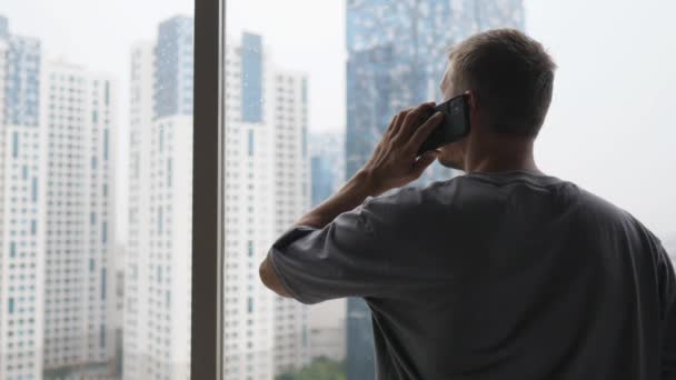 Achteraanzicht van een man die iemand belt terwijl hij uit het raam naar de wolkenkrabbers kijkt — Stockvideo