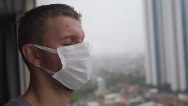 Лицо человека в маске, стоящего у окна с закрытыми глазами в знак истощения изоляции — стоковое видео